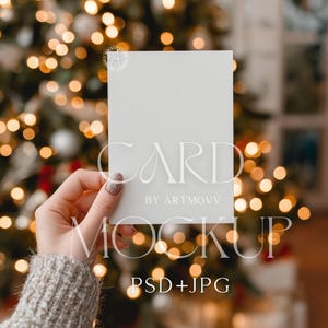 Könnte beinhalten: Ein weißes Karten-Mockup, das vor einem verschwommenen Weihnachtsbaum mit Bokeh-Lichtern gehalten wird. Die Karte trägt den Text "CARD BY ARTMOV" und "MOCKUP PSD+JPG" in einer stilvollen Schriftart. Der Hintergrund ist eine warme, festliche Szene.