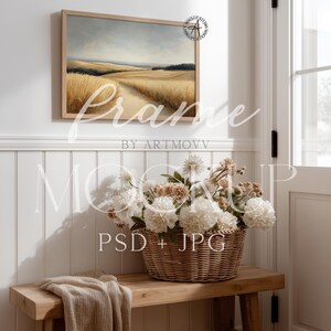 May include: A framed landscape painting with a wheat field scene hangs above a wooden bench. A woven basket filled with white and tan flowers sits on the bench. A beige throw blanket is draped on the bench.
