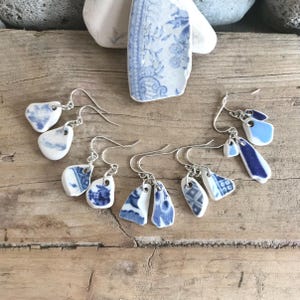 May include: A collection of handmade earrings featuring unique blue and white porcelain shards. The earrings showcase various shapes and patterns, including floral designs, with silver-colored ear wires. The pieces are arranged on a rustic wooden surface.