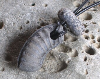 May include: A dark gray stone pendant with a ribbed texture, hanging from a black cord. The pendant is shaped like a curved, elongated oval.