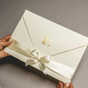 Op de afbeelding: Ivoorwitte uitnodigingsenvelop met een goudfolie monogram en een satijnen lint in een strik. De envelop wordt door handen tegen een neutrale achtergrond gehouden. Het monogram bevat de letters "IB".