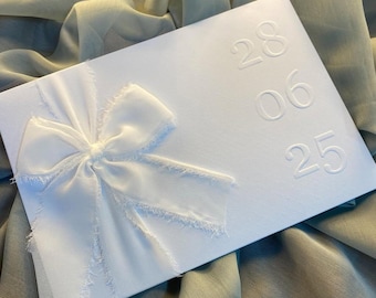 May include: A white invitation card with a large, textured bow on the front. The card has embossed numbers: 28, 06, and 25. The background is a soft, draped fabric.