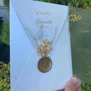 Op de afbeelding: Een witte huwelijksuitnodiging met gouden tekst "Emilia & Erivaldo 29.01.2025". De uitnodiging heeft gedroogde bloemen en een gouden zegel. Op de achtergrond is een groene uitnodiging met gouden bloemaccenten zichtbaar.