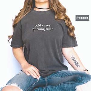 May include: A dark gray t-shirt with the text "cold cases burning truth" printed in white on the front. The shirt is being worn by a person with long brown hair.