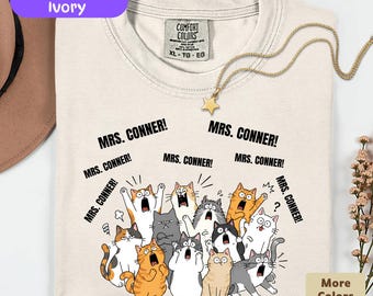 May include: Ivory Comfort Colors t-shirt featuring a graphic of cartoon cats with the text "MRS. CONNER!". The cats are in various poses, with open mouths and surprised expressions. A gold necklace with a star pendant is draped on the shirt.