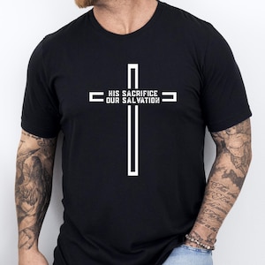 May include: Black t-shirt featuring a white cross design. The cross has the text "HIS SACRIFICE OUR SALVATION" printed across the horizontal bar. The shirt is a crew neck style and appears to be made of a soft material.
