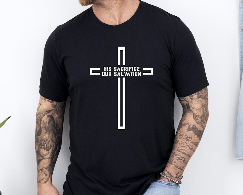 May include: Black t-shirt featuring a white cross design. The cross has the text "HIS SACRIFICE OUR SALVATION" printed across the horizontal bar. The shirt is a crew neck style and appears to be made of a soft material.