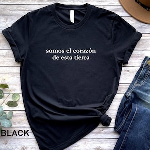 May include: Black t-shirt with the Spanish text "somos el corazón de esta tierra" in white. The shirt is paired with blue jeans and a brown hat. The word "BLACK" is printed at the bottom left.