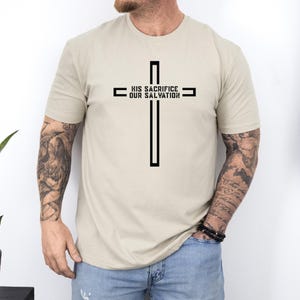 May include: Beige t-shirt featuring a black cross design with the text "HIS SACRIFICE OUR SALVATION". The graphic is centered on the front of the shirt. The shirt is made of a soft, neutral-colored fabric.