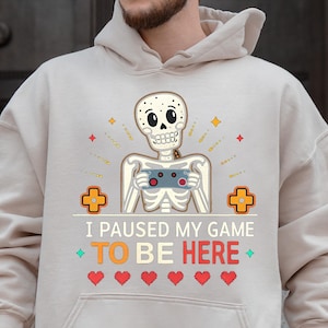 A man wearing a sand hoodie with the words “i paused my game to be here” and a cute skeleton smiling and holding a game console