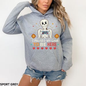 A woman wearing a grey hoodie with the words “i paused my game to be here” and a cute skeleton smiling and holding a game console