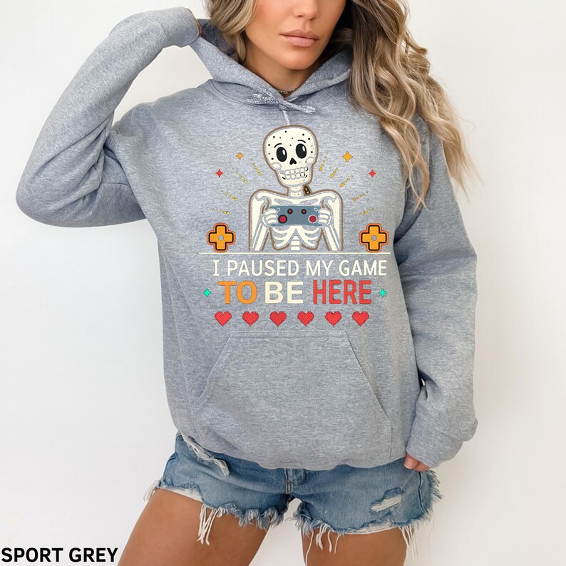 A woman wearing a grey hoodie with the words “i paused my game to be here” and a cute skeleton smiling and holding a game console