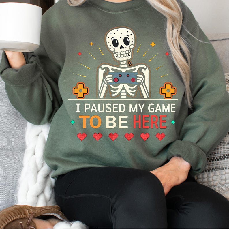 A woman wearing a green sweatshirt with the words “i paused my game to be here” and a cute skeleton smiling and holding a game console