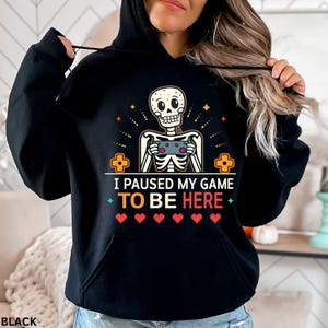 A woman wearing a black hoodie with the words “i paused my game to be here” and a cute skeleton smiling and holding a game console