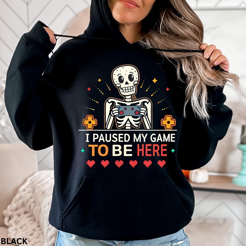 A woman wearing a black hoodie with the words “i paused my game to be here” and a cute skeleton smiling and holding a game console