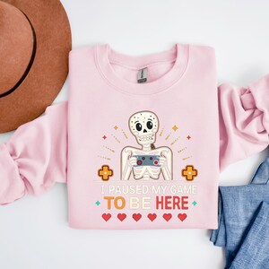 a pink sweatshirt with the words “i paused my game to be here” and a cute skeleton smiling and holding a game console
