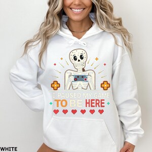 A woman wearing a white hoodie with the words “i paused my game to be here” and a cute skeleton smiling and holding a game console