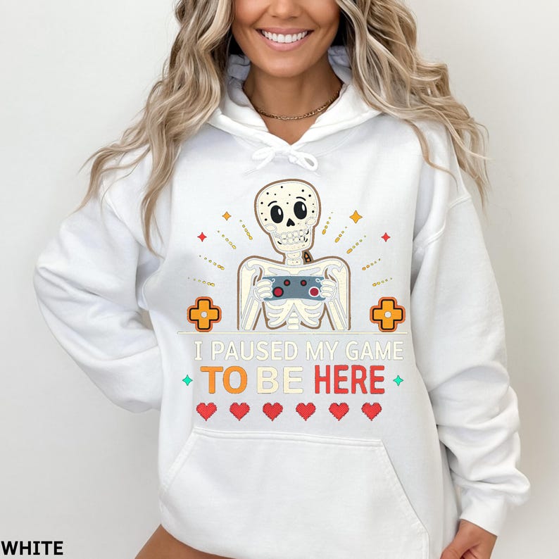 A woman wearing a white hoodie with the words “i paused my game to be here” and a cute skeleton smiling and holding a game console