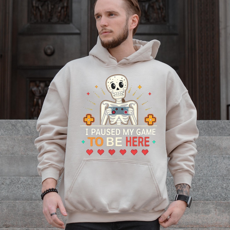 A man wearing a sand hoodie with the words “i paused my game to be here” and a cute skeleton smiling and holding a game console