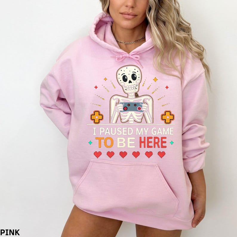 A woman wearing a pink hoodie with the words “i paused my game to be here” and a cute skeleton smiling and holding a game console