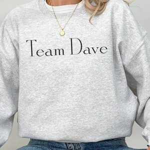 May include: A grey sweatshirt with the text "Team Dave" printed on the front. The model is wearing the sweatshirt with blue jeans and black trainers.