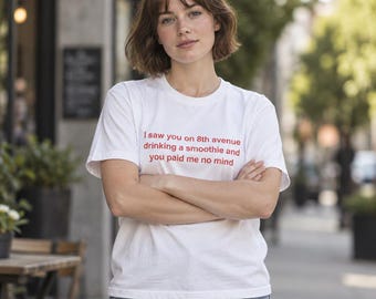 May include: A white short-sleeved t-shirt featuring red text printed on the chest that reads: I saw you on 8th avenue drinking a smoothie and you paid me no mind. The shirt has a crew neckline and a standard fit, paired with blue denim jeans.