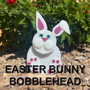Puede incluir: Un conejito de Pascua de vidrio blanco, en forma de bobblehead, con orejas rosas y ojos azules. El conejito está sentado sobre una base de madera. "Easter Bunny Bobblehead" está escrito en texto negro debajo del conejito.