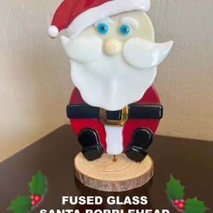 May include: Fused glass Santa bobblehead with a red hat, white beard, and black boots. The bobblehead is standing on a wooden base. "Fused Glass Santa Bobblehead with Pam Jacobson" is written below the base.