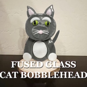 Puede incluir: Un cabezón de gato de vidrio fundido gris y blanco con ojos verdes. El gato está sentado sobre una base de madera.