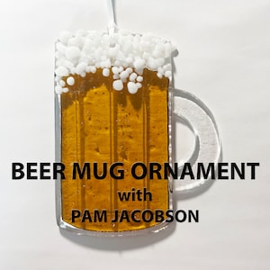 May include: A glass beer mug ornament with a white foam top and a clear handle. The mug is filled with a golden yellow liquid. The text "BEER MUG ORNAMENT with PAM JACOBSON" is printed below the mug.