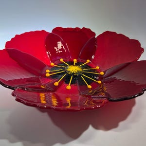 May include: A red glass flower bowl with a black and yellow center. The bowl is shaped like a poppy flower with multiple petals.
