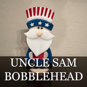 May include: A glass Uncle Sam bobblehead figurine with a red, white, and blue hat and beard. The figurine is standing on a wooden base. "Uncle Sam Bobblehead" is written in white text below the figurine.