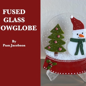 May include: Fused glass snowglobe featuring a snowman with a red hat and green scarf, and a green Christmas tree with red candy cane ornaments. The snowglobe has a red base with white polka dots and the text "Fused Glass Snowglobe" and "By Pam Jacobson".