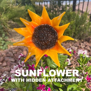 May include: A glass sunflower with yellow petals and an orange center. The sunflower is in a garden setting with green leaves and pink flowers.