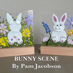 May include: Two glass bunny scene decorations. Each features a white bunny with pink accents, surrounded by colorful flowers and greenery. The text "BUNNY SCENE By Pam Jacobson" is at the bottom. The decorations sit on wooden bases.