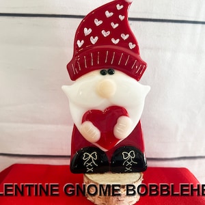 May include: A red glass gnome bobblehead holding a red heart. The gnome is wearing a red hat with white hearts and black boots.