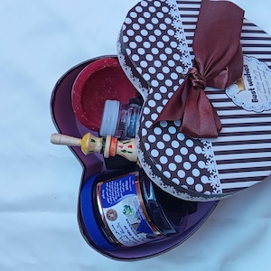 Può includere: Una scatola regalo a forma di cuore con un motivo a righe marroni e bianche e pois bianchi. La scatola è legata con un nastro marrone e ha le parole "Auguri" stampate su di essa. La scatola è aperta e contiene un piccolo cucchiaio di legno, un piccolo barattolo di liquido blu e una piccola ciotola rossa.