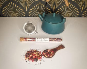 May include: A teal ceramic teapot with a woven handle, a metal tea infuser, and a glass tube of "Berry Spice" tea. A wooden spoon and a pile of loose tea are also present, suggesting a tea set.