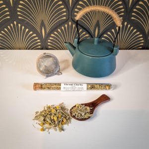 Op de afbeelding: Een teal keramische theepot met een touw handvat staat naast een roestvrijstalen thee-ei en een glazen buisje met het opschrift "Throat Clarity". Een houten lepel bevat losse theebladeren.