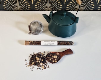 May include: A teal ceramic teapot with a woven handle is next to a stainless steel tea infuser and a glass tube of tea. A wooden spoon holds loose tea leaves, with a pile of tea leaves beside it. The tube reads "Rooibos Earl".
