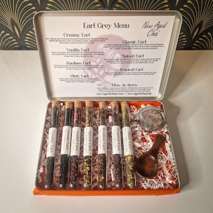 May include: An open metal tin with an "Earl Grey Menu" printed on the lid. Inside are glass tubes filled with tea, a small wooden scoop, and a tea strainer. The tea varieties include Creamy, Classic, Vanilla, Spiced, Rooibos, Tropical, and Minty Earl.