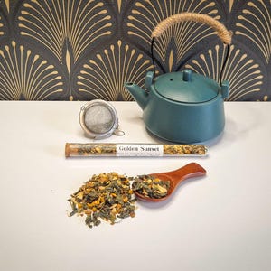 May include: A teal ceramic teapot with a woven handle, a stainless steel tea infuser, a glass tube of tea labelled "Golden Sunset", and a wooden spoon filled with loose tea. The tea is a mix of green and yellow herbs.