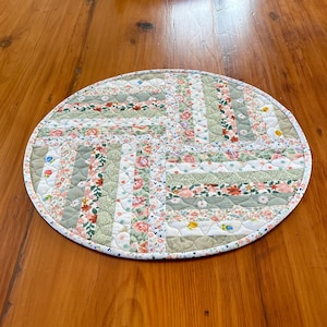 May include: A circular, quilted placemat with a floral pattern in pink, green, and white. The placemat has a patchwork design and a white border. It is placed on a wooden table.