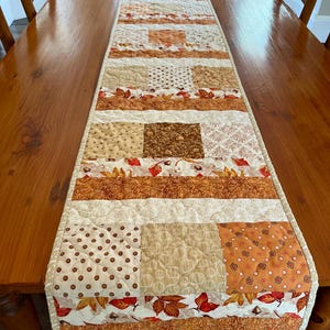 May include: A patchwork table runner in autumnal colours. The runner is made up of squares and strips of fabric in cream, beige, brown, and orange. Some squares have polka dots, pumpkin patterns, or leaf designs. It is placed on a wooden table.