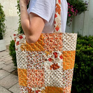 Peut inclure: Un sac fourre-tout matelassé présentant un motif patchwork composé de divers tissus aux couleurs automnales. Les carrés affichent des motifs de feuilles orange, de petites fleurs et de pois marron sur fond crème. Le sac possède deux anses assorties.