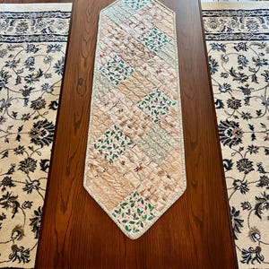 May include: A diamond-shaped quilted table runner featuring a patchwork design of beige fabric, green holly leaves with red berries, and brown deer silhouettes. The runner has a light-coloured border and is stitched with a textured, wavy pattern.