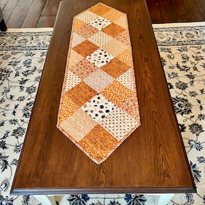 May include: A quilted table runner featuring a diamond-shaped patchwork design in shades of orange, yellow, and white. The fabric squares display various patterns including autumn leaves, polka dots, and floral motifs with a pointed end finish.