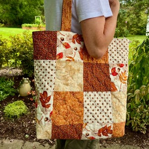 May include: A quilted tote bag with a patchwork design featuring autumn leaves and polka dots in shades of brown, cream, and orange. The bag has a matching patterned strap and is carried over the shoulder.