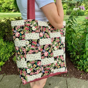 May include: A quilted tote bag with a floral pattern featuring pink, white, and red roses on a black background. The bag has a red and white patterned strap and trim. The bag is held by a person.
