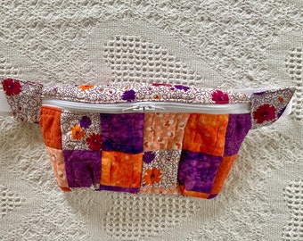 May include: A patchwork fanny pack with a white zipper and a white strap. The fabric is a mix of orange, purple, and white floral patterns.